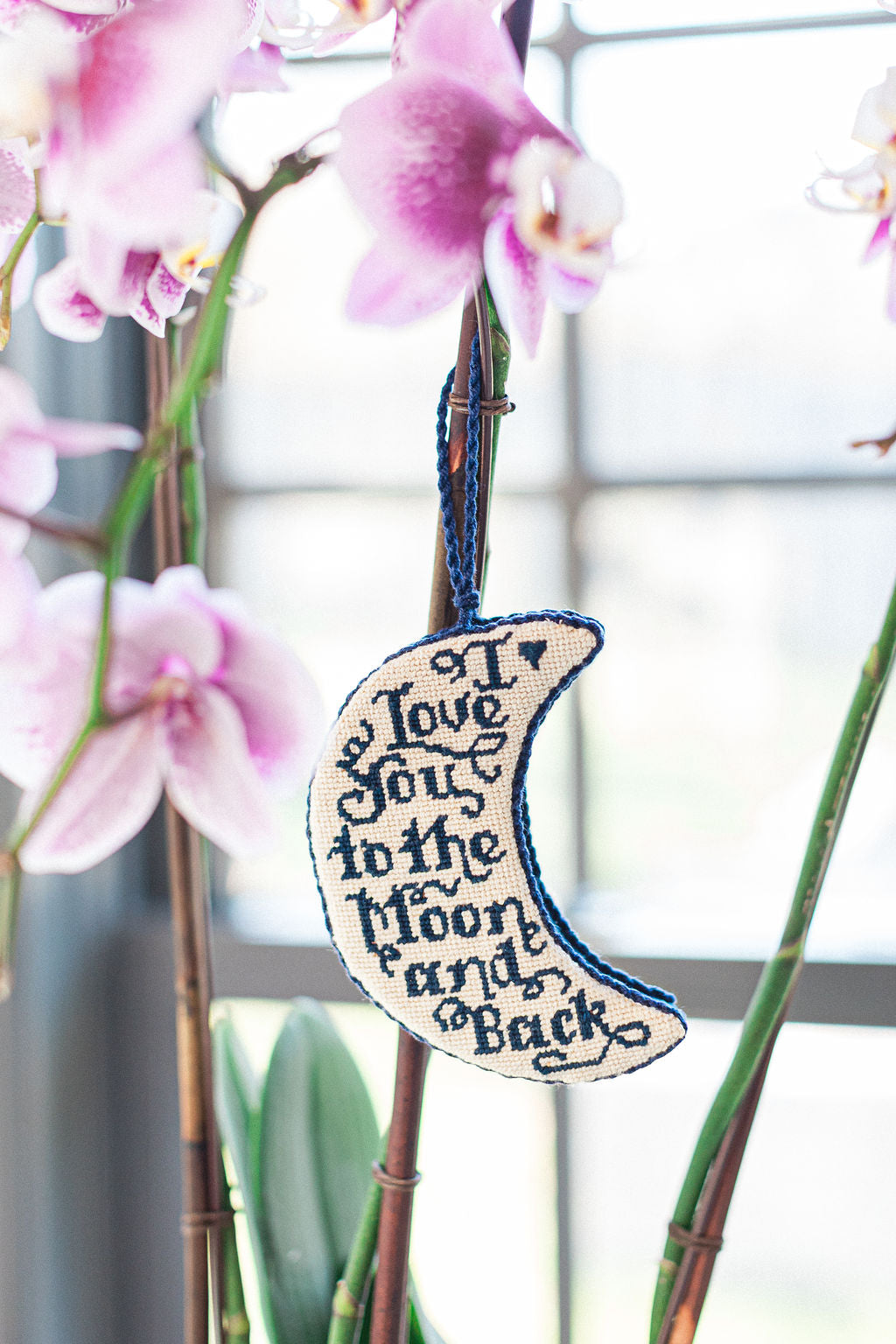 Decorative needlepoint moon-shaped sign with 'I love you to the moon and back' text, surrounded by pink orchids. | The Pepper Pot, Barrington, IL