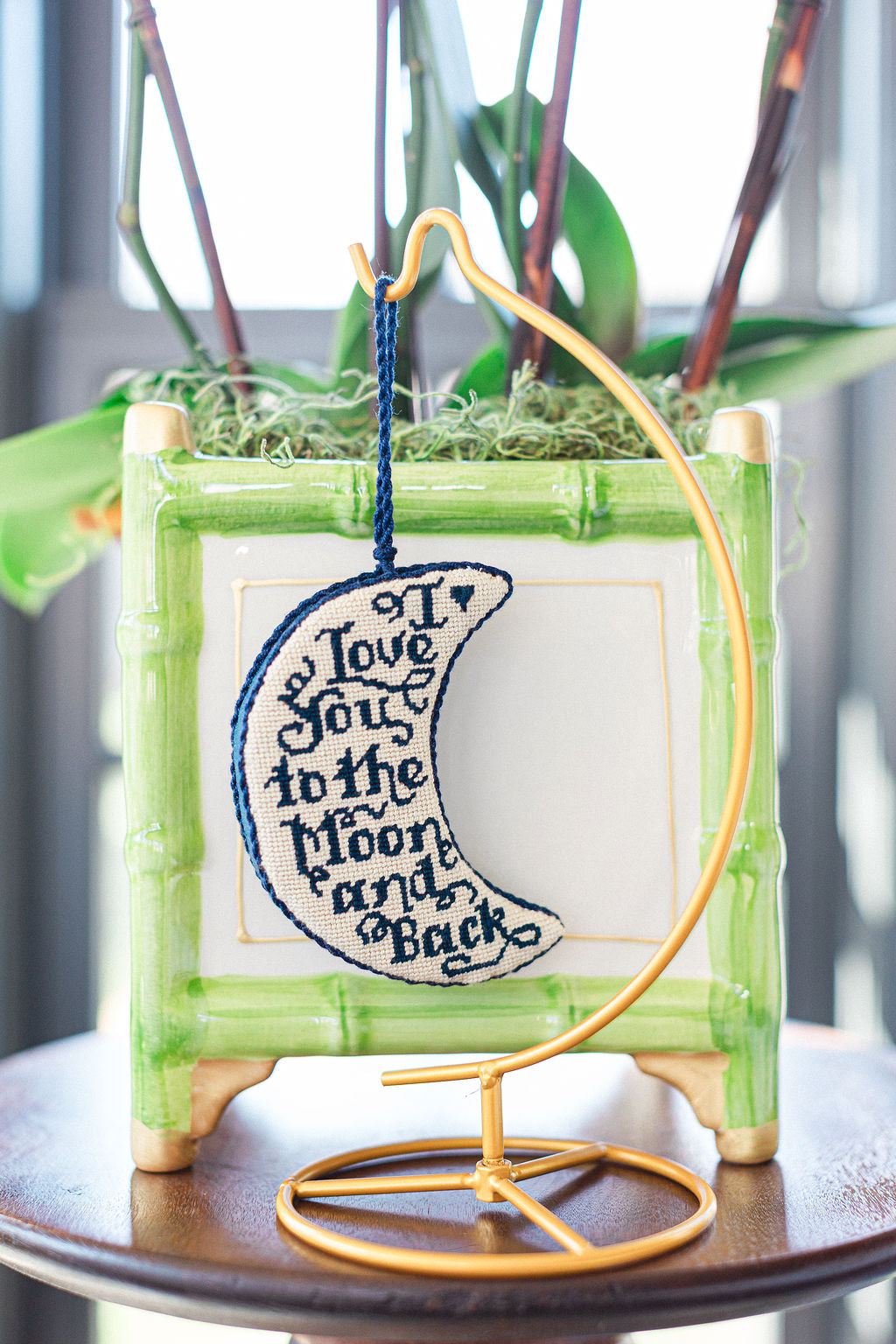 Decorative needlepoint ornament  with a crescent moon design and text, placed on a surface with a blurred background.  | The Pepper Pot Barrington, IL 