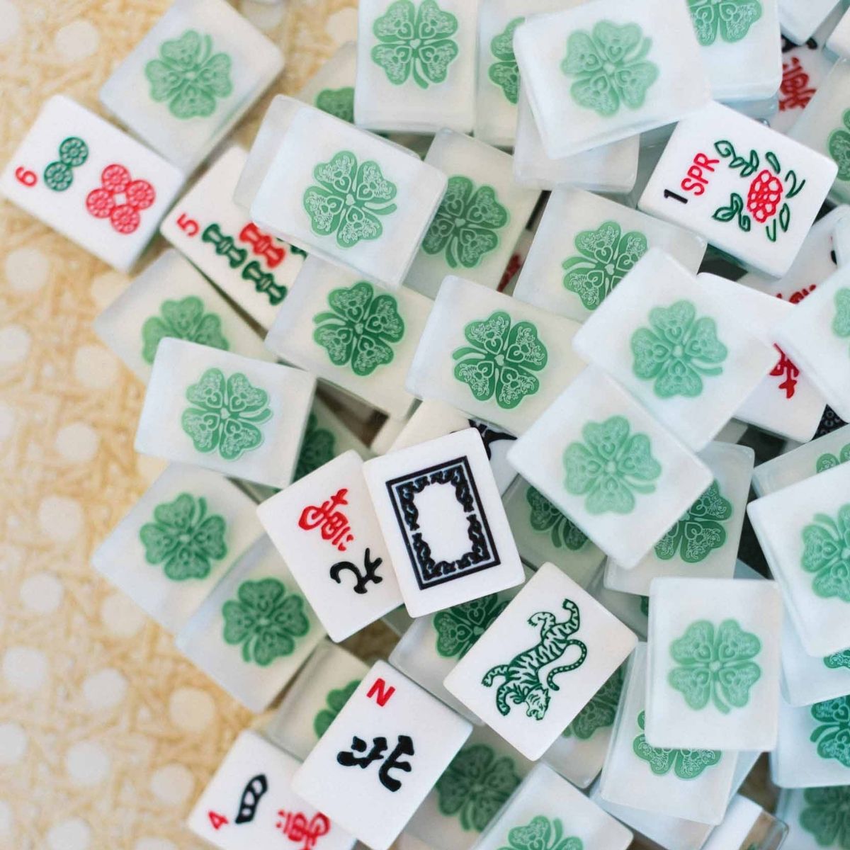 A pile of Chinese mahjong tiles on a light beige patterned surface, showing white tiles with green floral designs and red, black, and green scoring and character markings.