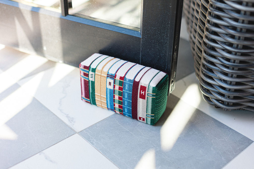 Colorful needlepoint door stopper with a book design on a checkered floor with a wicker basket in the background. 