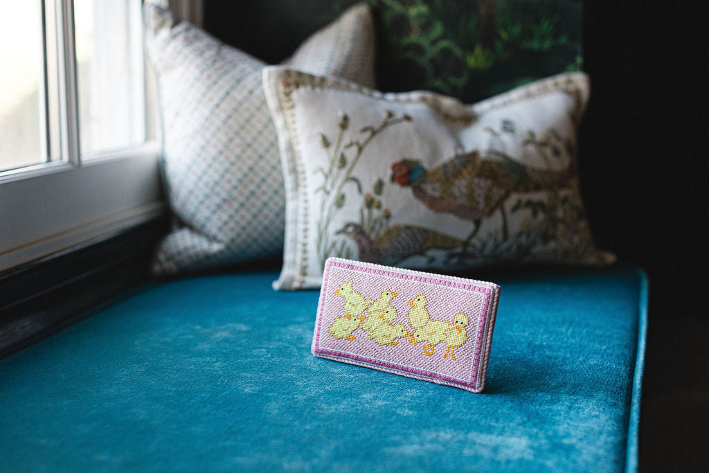 Small pink needlepoint  card with duck illustrations on a blue surface with decorative pillows in the background.  | The Pepper Pot Barrington, IL 