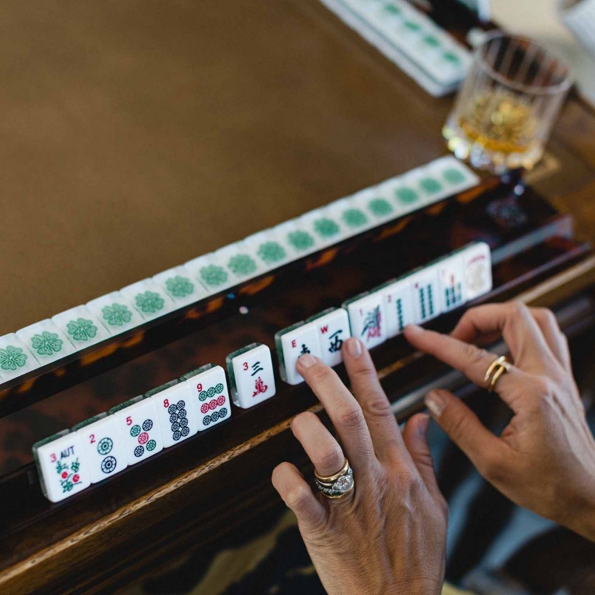 A close-up of a mahjong game in progress on a wooden table, with a person's hands holding and selecting tiles. A row of green-backed tiles is visible in the background, along with a glass of amber liquid.