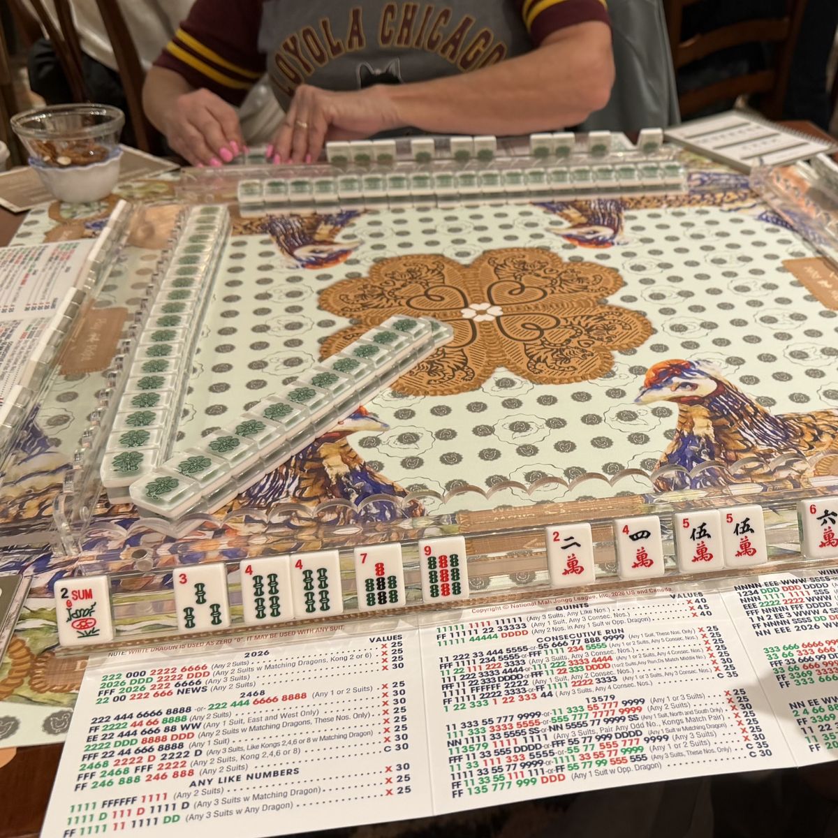 Mahjong game set on a table with a person partially visible above the board at The Pepper Pot in Barrington, IL