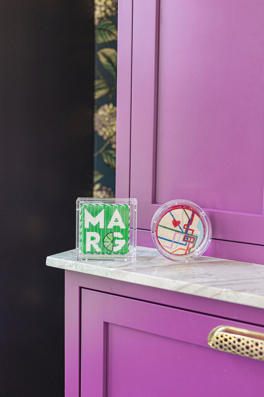 Two finished acrylic needlepoint coasters  on a marble surface in front of a purple cabinet.  | The Pepper Pot Barrington, IL 