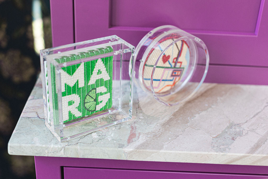 Decorative acrylic pieces with finished needlepoint projects on a marble surface with a purple cabinet in the background. | The Pepper Pot, Barrington, IL