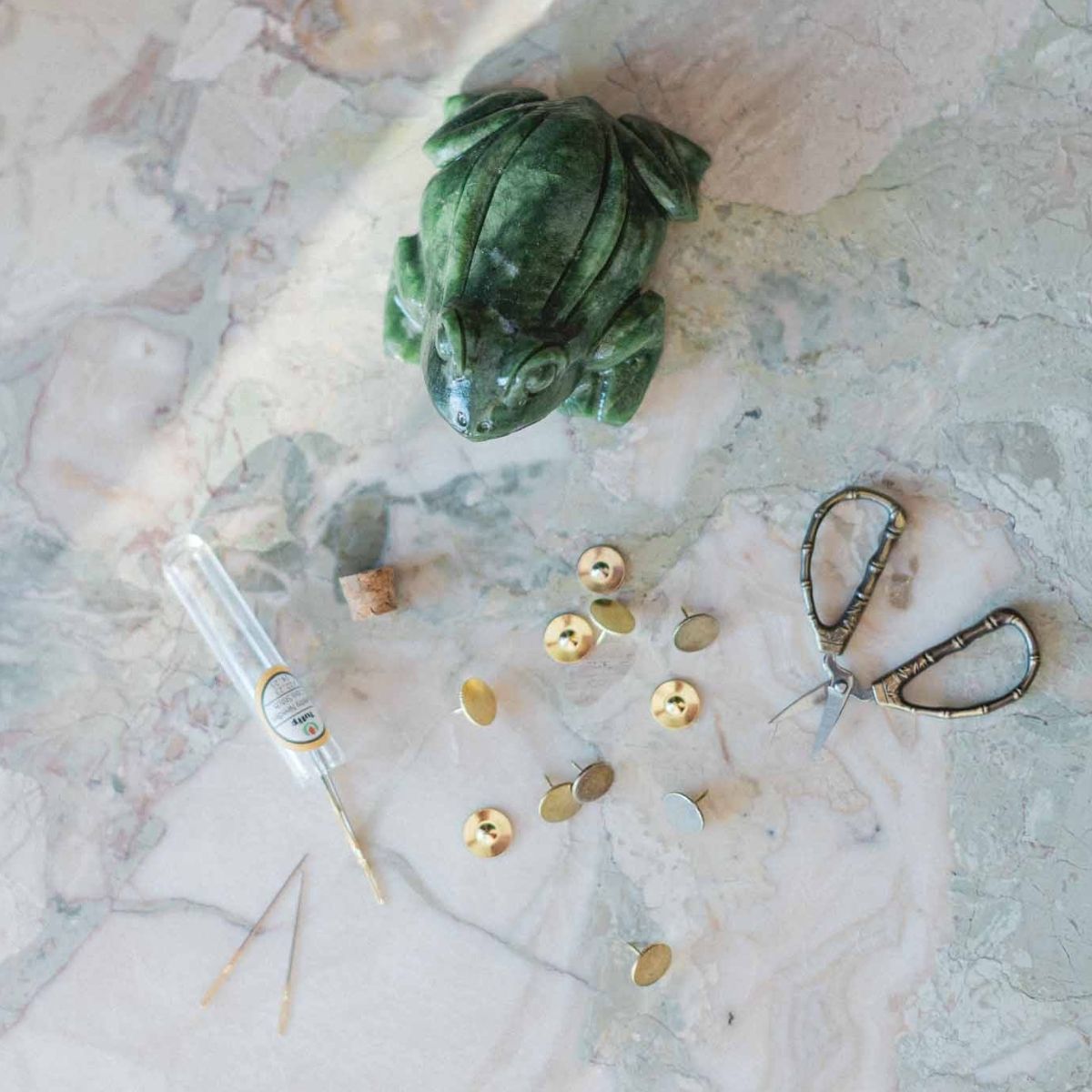 Green stone frog, gold beads, and scissors on a marble surface. Needlepoint supplies and accessories available at The Pepper Pot in Barrington, IL