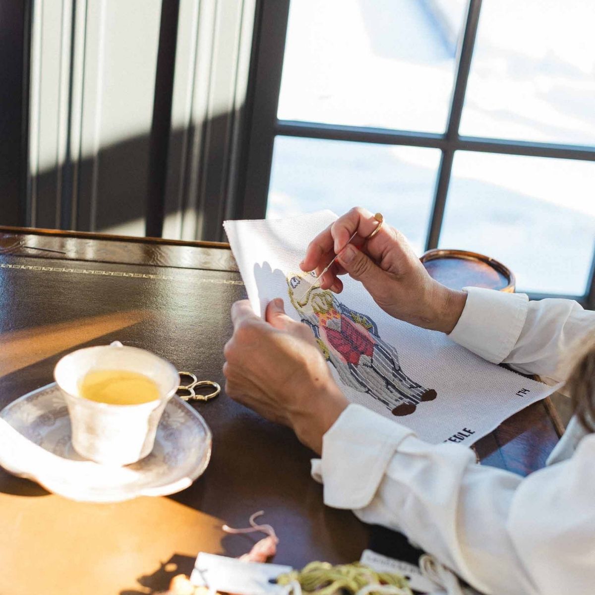 Person holding a white needlepoint canvas with a cartoon frog design on a table with a cup and saucer. 