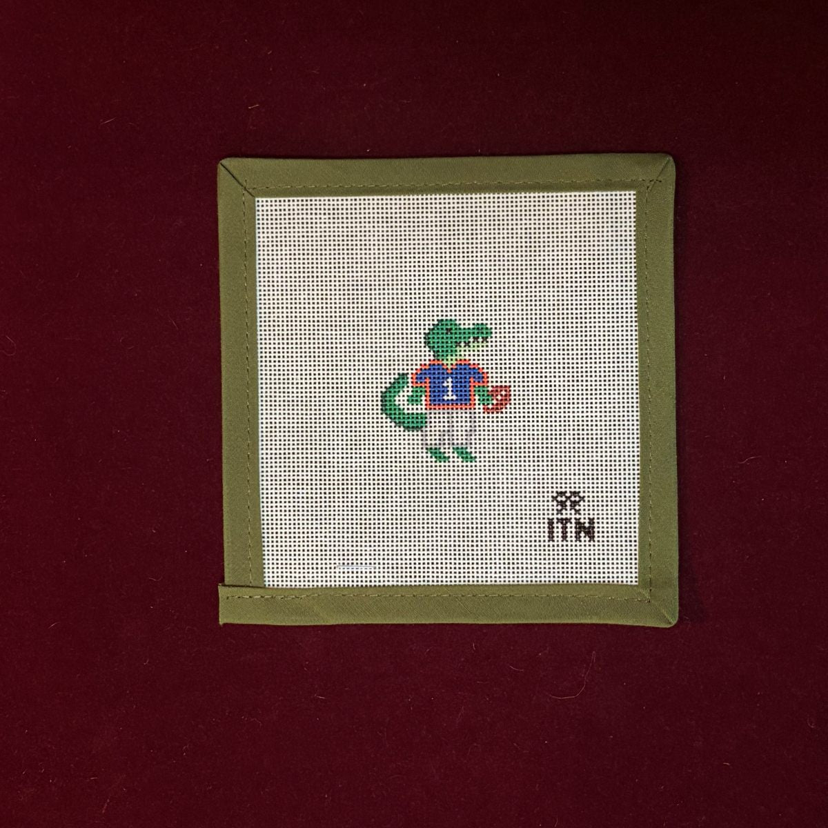 Needlepoint canvas design, gator character on fabric with a green border, on a red background.