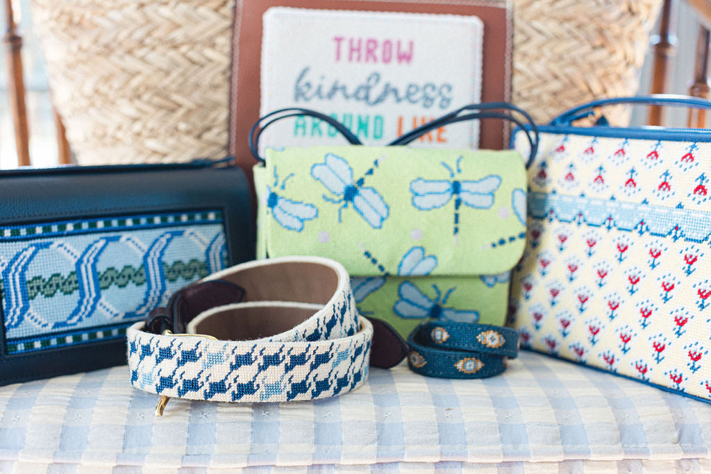 Collection of needlepoint bags and accessories on a checkered surface with a sign in the background. | The Pepper Pot, Barrington, IL