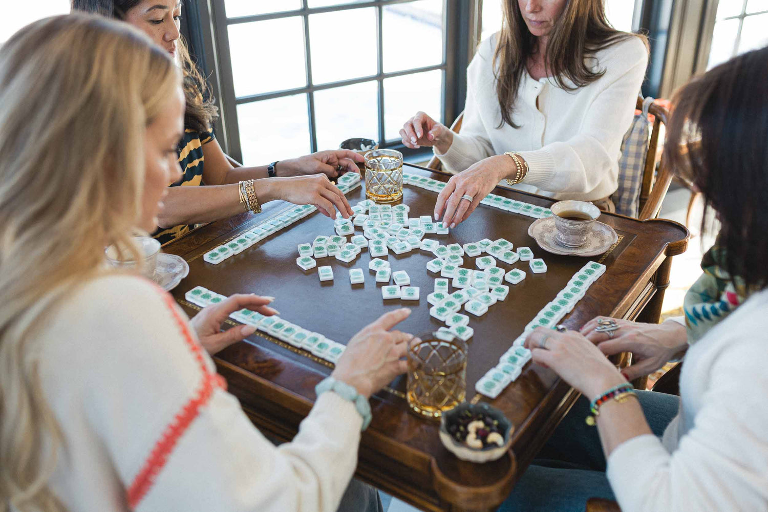 Four women playing a game of mahjong around a wooden table with drinks and snacks. The Pepper Pot | Needlepoint and Mahjong Specialty Store in Barrington, IL