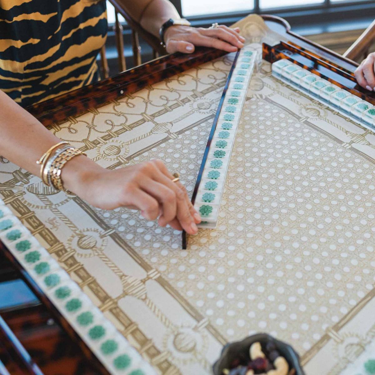 Close-up of a person's hands on a decorative mahjong mat with intricate patterns and a tray of mahjong tiles. 