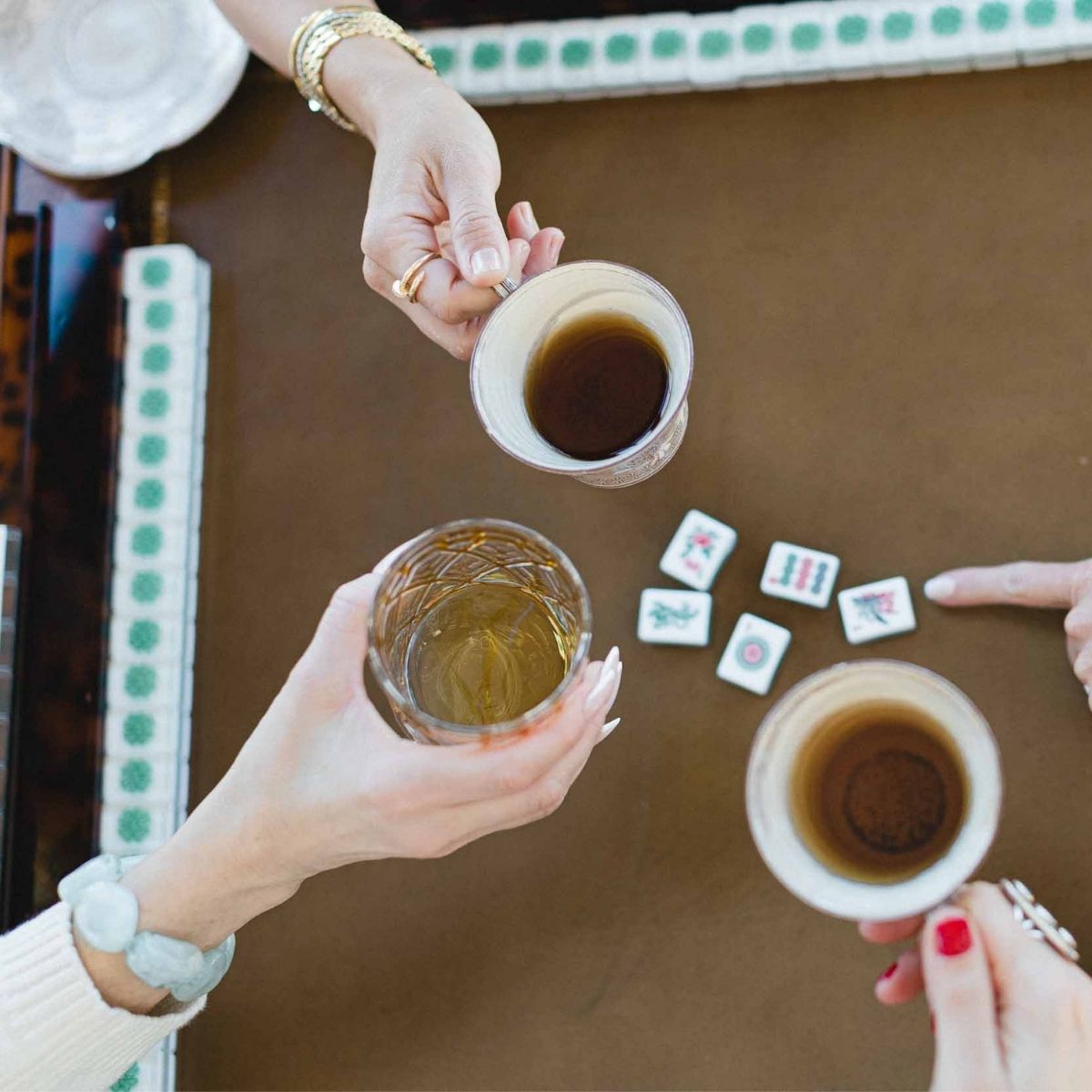 Three hands holding clear drinking glasses filled with dark tea over a brown mahjong table, with small tiles scattered nearby.