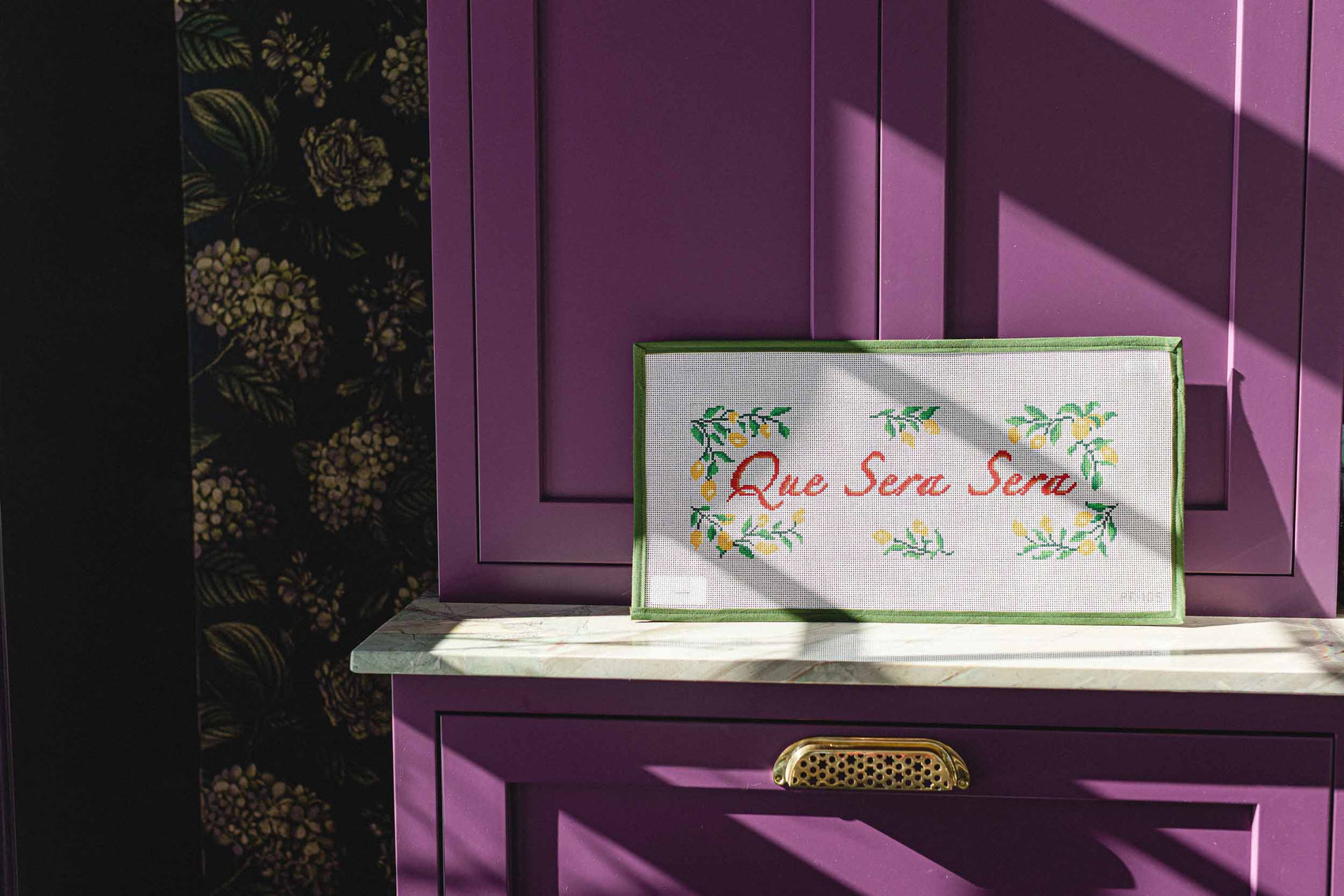 Decorative sign with 'Que Sera Sera' on a purple cabinet with floral wallpaper. The Pepper Pot | Needlepoint and Mahjong Specialty Store in Barrington, IL