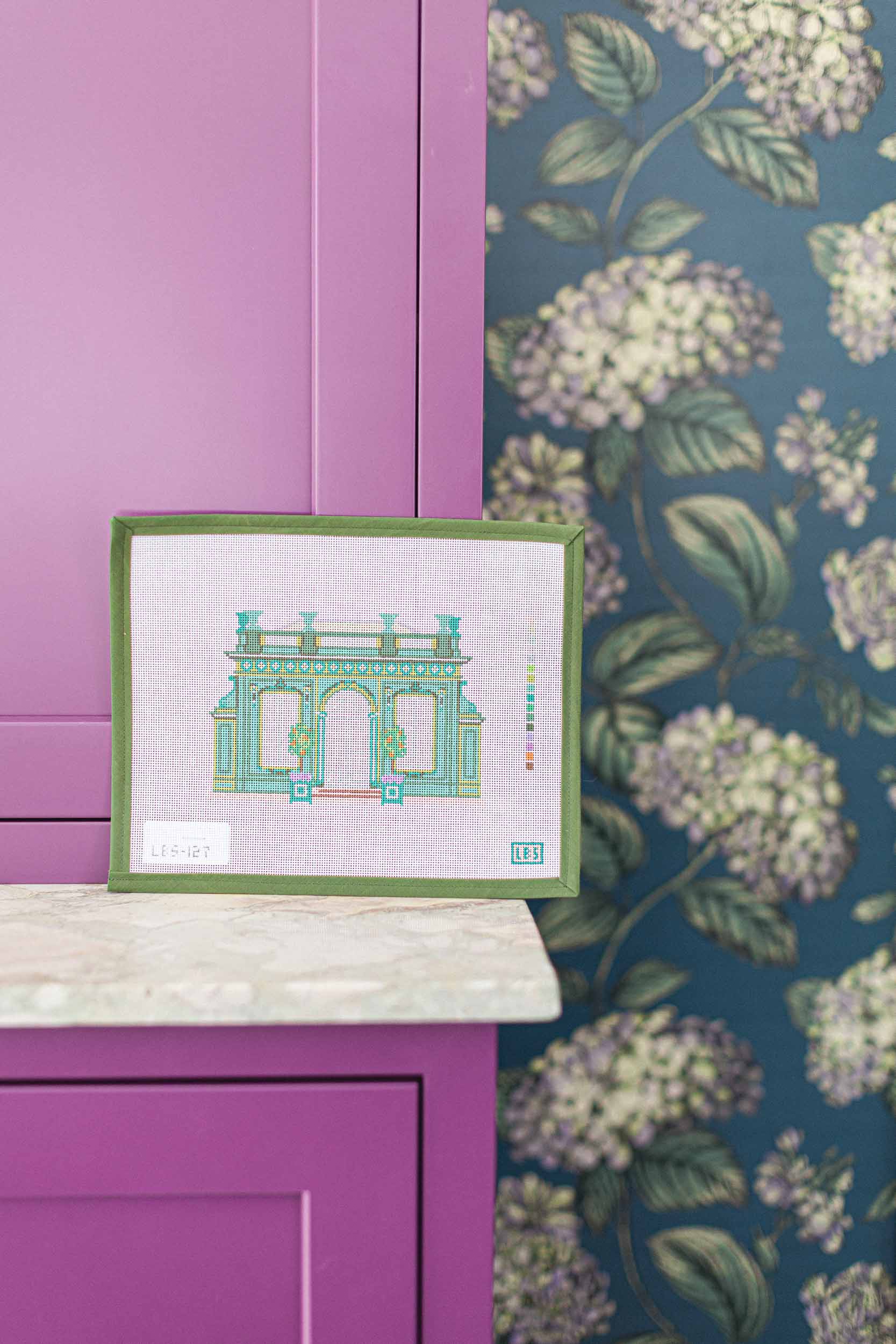 Canvas needlepoint artwork on a purple surface with floral wallpaper in the background.  | The Pepper Pot Barrington, IL 