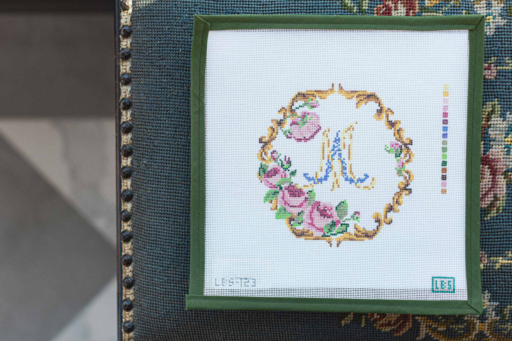 Needlepoint or Cross-stitched letter 'M' with floral design in a green frame on a textured fabric background.  | The Pepper Pot Barrington, IL 