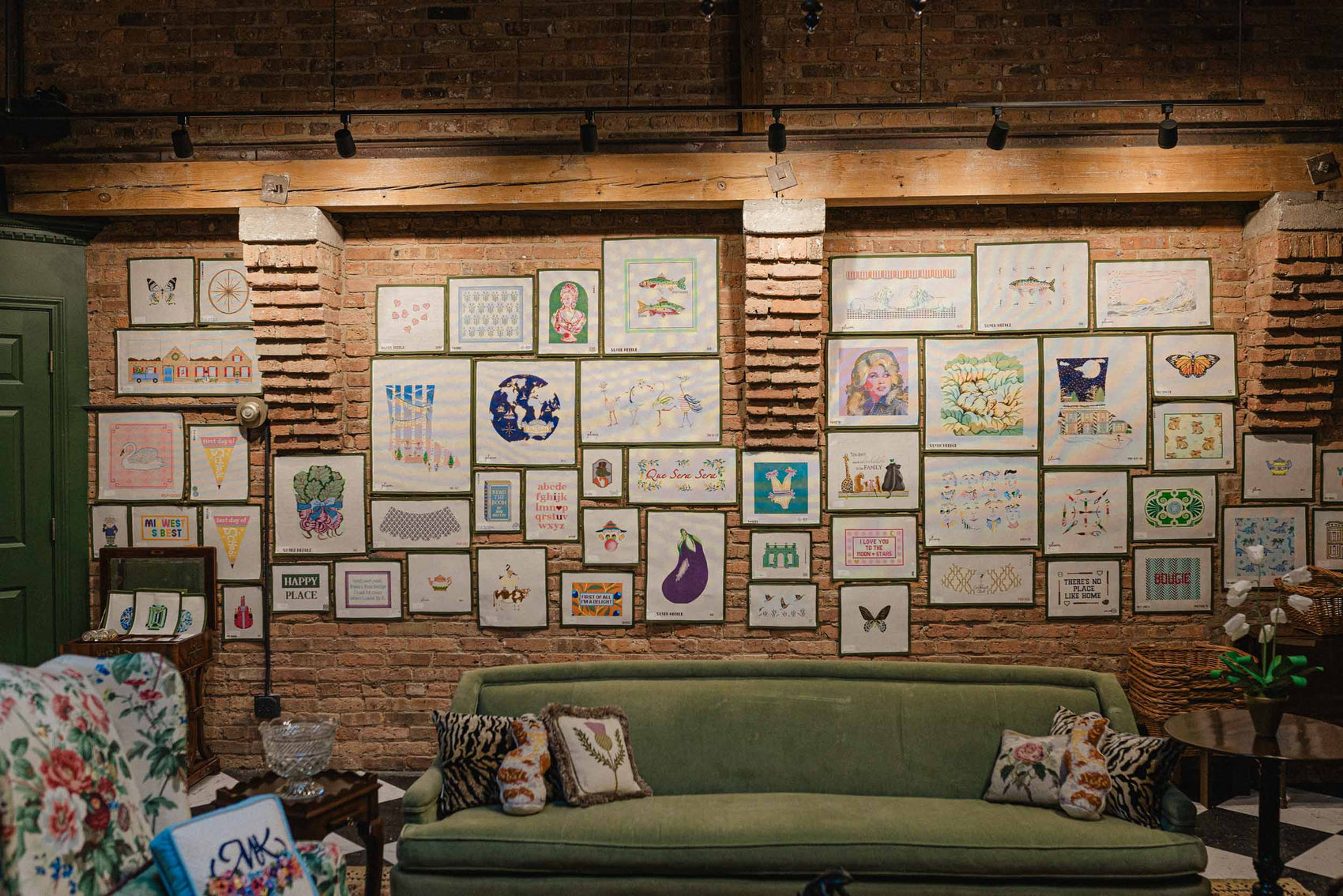 Green sofa in a room with a wall displaying various hand painted needlepoint canvas options available at The Pepper Pot | Needlepoint and Mahjong Specialty Store in Barrington, IL