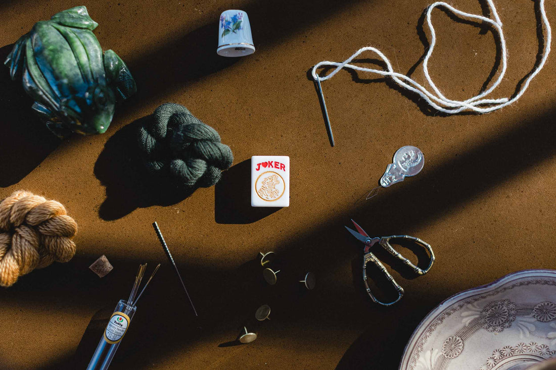 Yarn, scissors, needlepoint and cross stitch supplies  and a mahjong joker tile on a brown surface. | The Pepper Pot Barrington, IL 