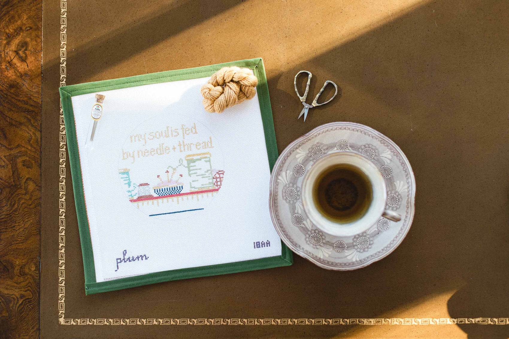 Teacup and saucer with a handp painted needlepoint canvas on a wooden surface. Canvas has text that reads 'my soul is fed by needle and thread'. The Pepper Pot | Needlepoint and Mahjong Specialty Store in Barrington, IL