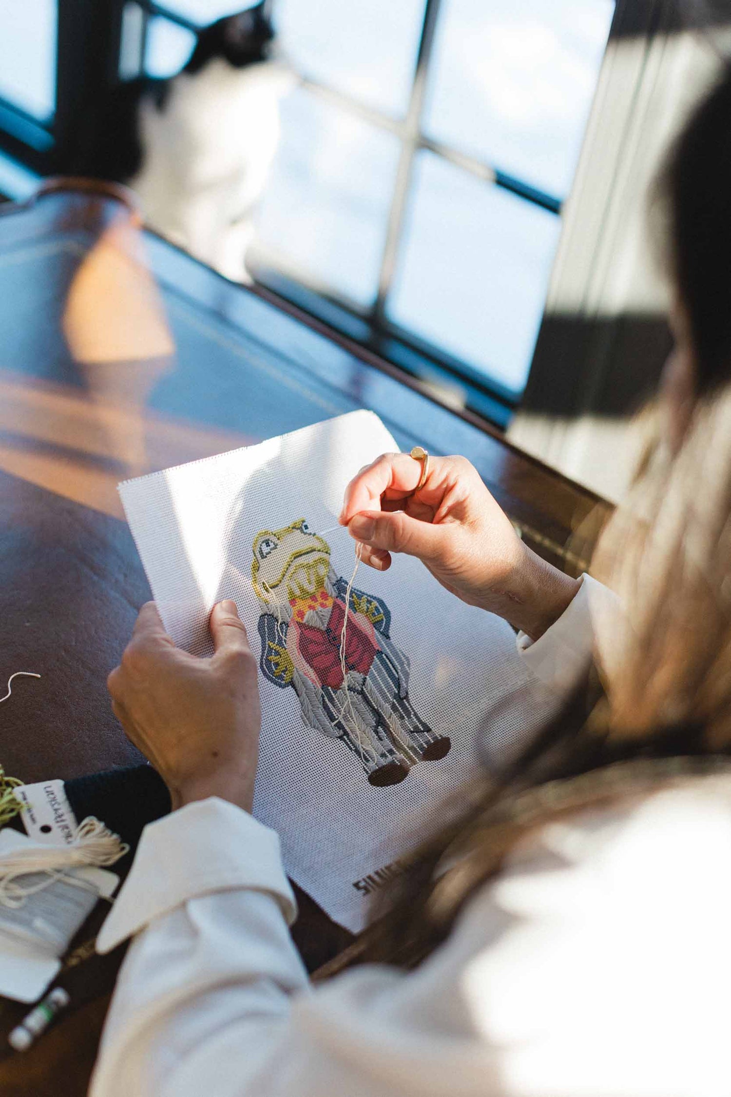 Person holding a hand painted needlepoint canvas with a colorful frog design, possibly a cartoon character, in a blurred indoor setting. | The Pepper Pot, Barrington, IL