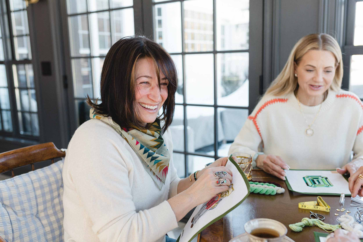 Two women sitting at a table with needlepoint and cross stitch craft materials, smiling and engaged in an activity. | The Pepper Pot, Barrington, IL