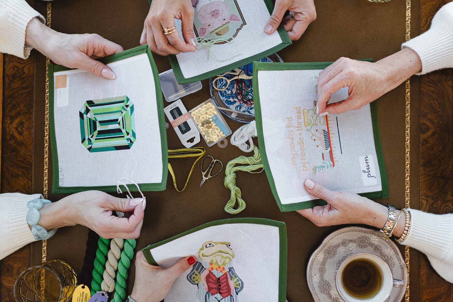 People working on embroidery needlepoint projects with hand painted needlepoint canvases, materials and a cup of tea on a wooden table. | The Pepper Pot, Barrington, IL