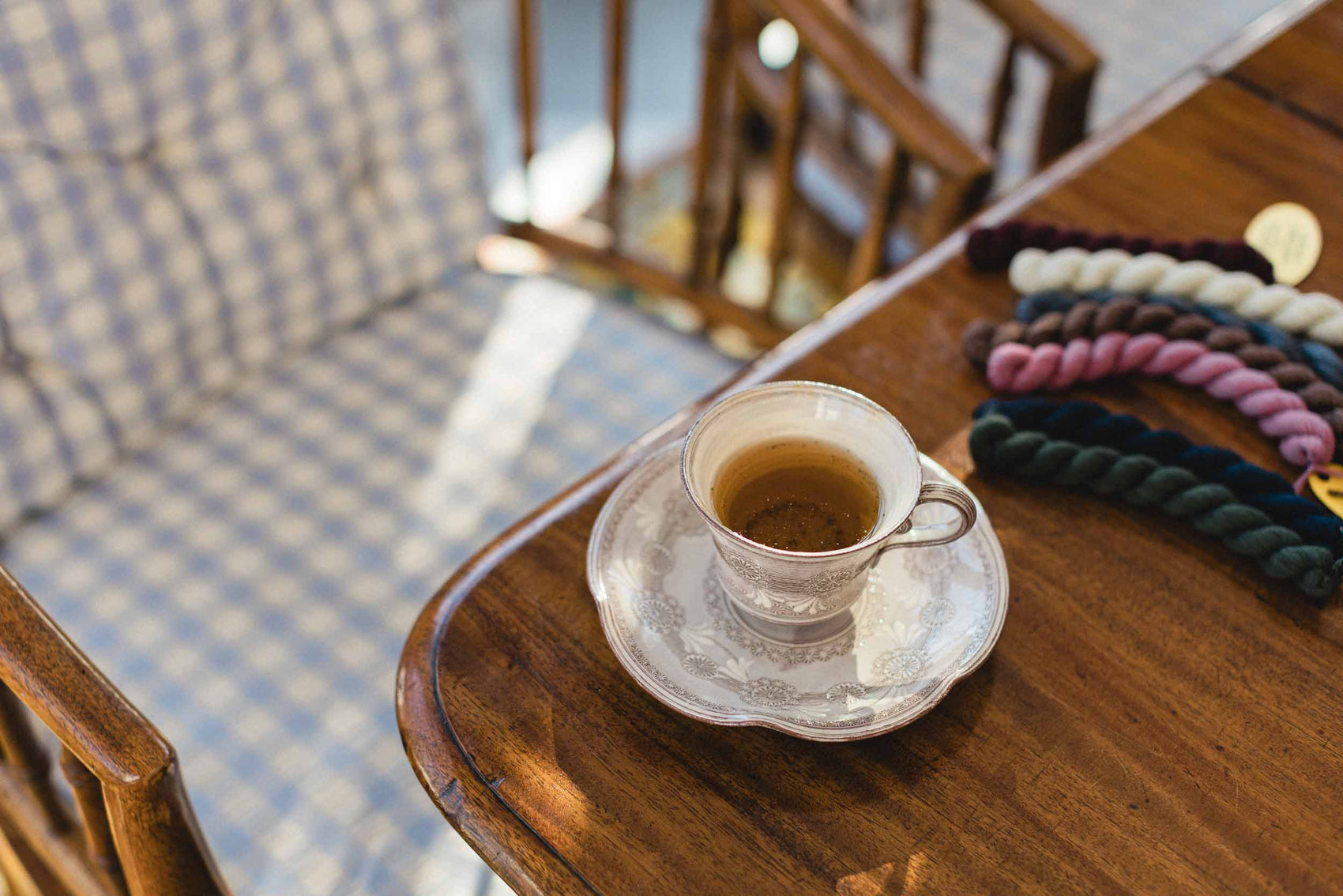 Teacup on a wooden table with a checkered chair in the background. The Pepper Pot | Needlepoint and Mahjong Specialty Store in Barrington, IL