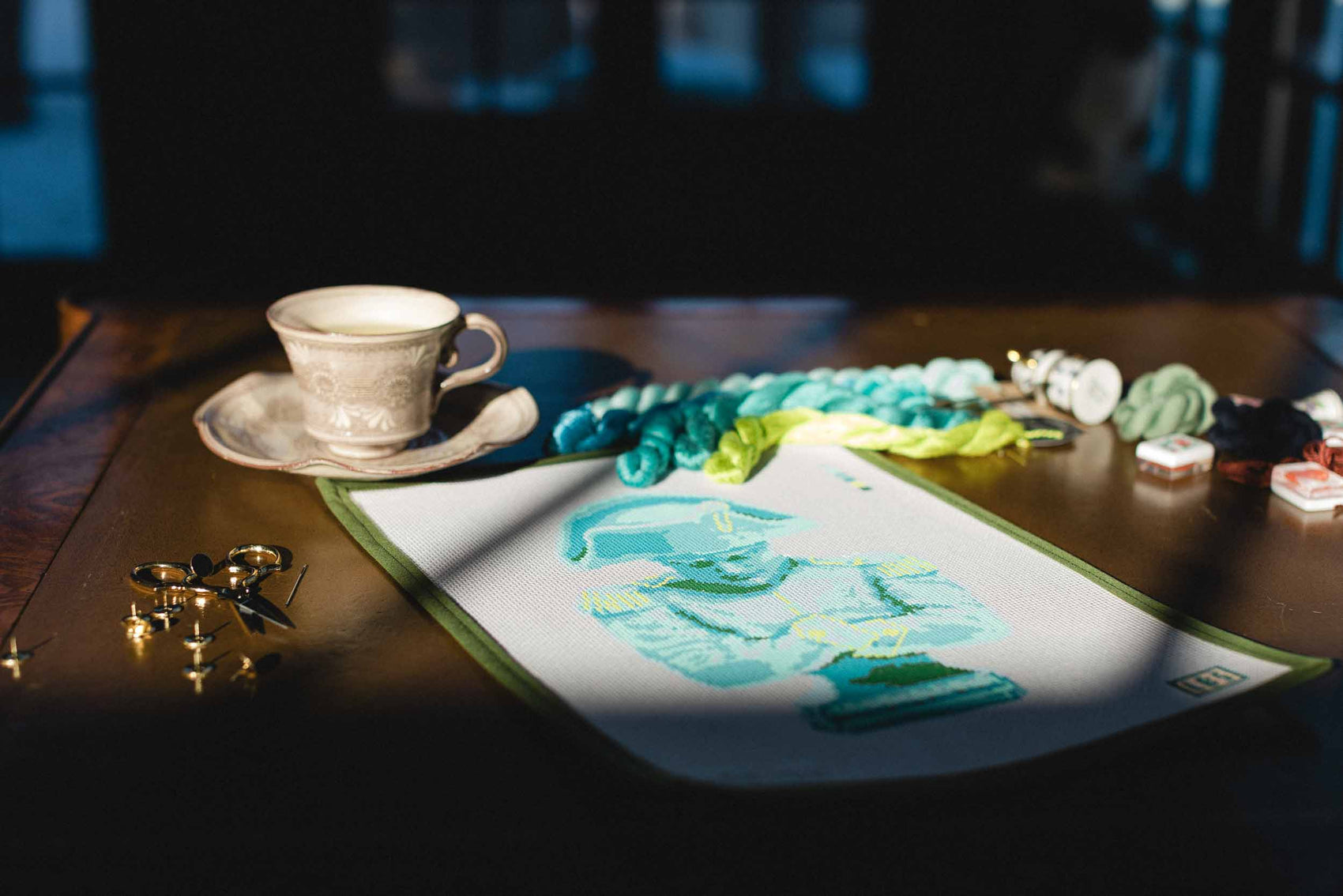 Cup and saucer on a table with needlepoint art supplies and a piece of canvas with blue bust design. | The Pepper Pot, Barrington, IL