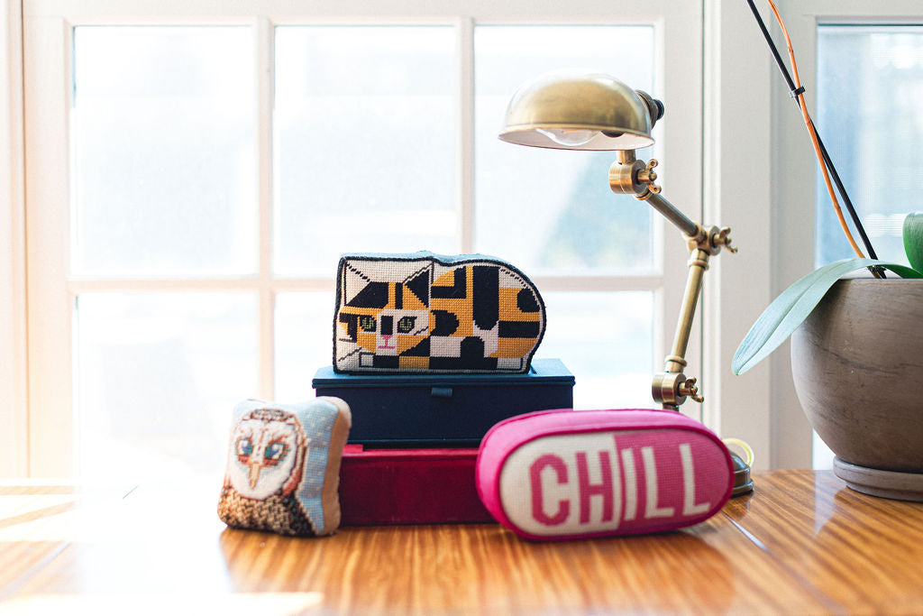 Decorative needlepoint  items including a colorful pouch, owl-themed pillow, and 'CHILL' pillow on a wooden surface with a lamp and plant in the background. | The Pepper Pot Barrington, IL 