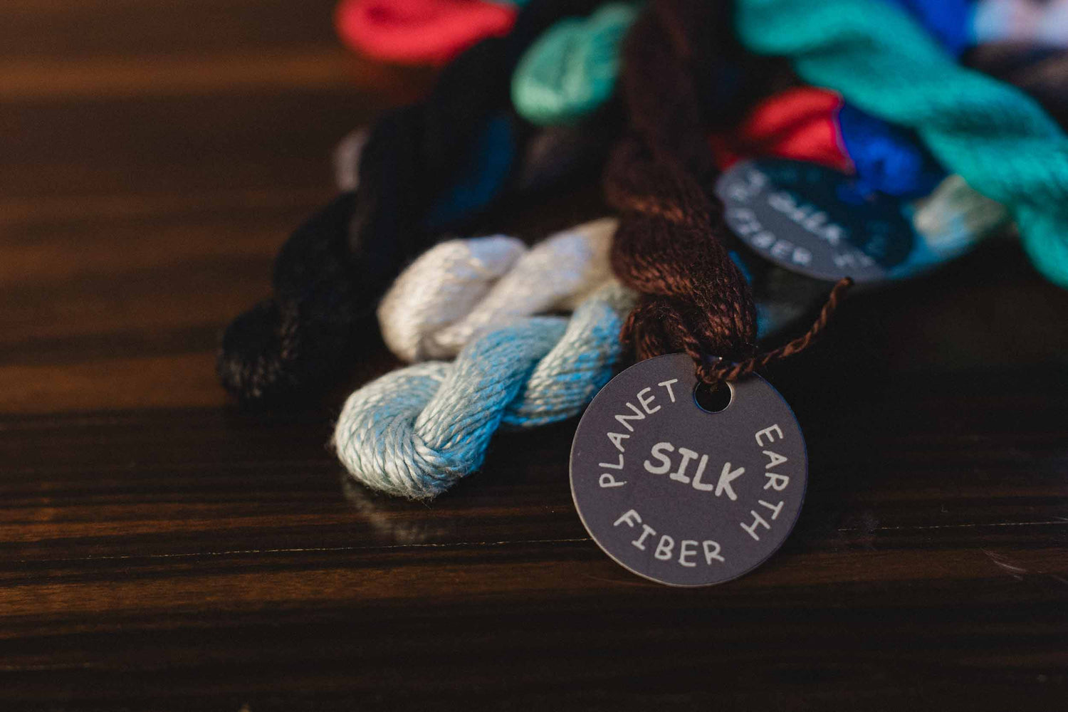 Colorful skeins of yarn with a 'Planet Silk Fiber' tag on a wooden surface.  | The Pepper Pot, Needlepoint and Mahjong Specialty Store | Barrington, IL