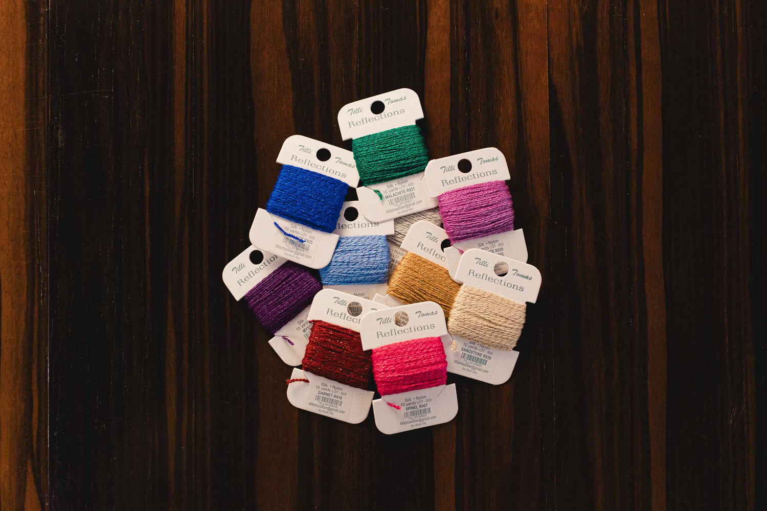 Colorful Tilli Tomas brand Reflections Threads and Fibers for needlepoint canvases. Ccards displayed against a wooden background. | The Pepper Pot, Barrington, IL