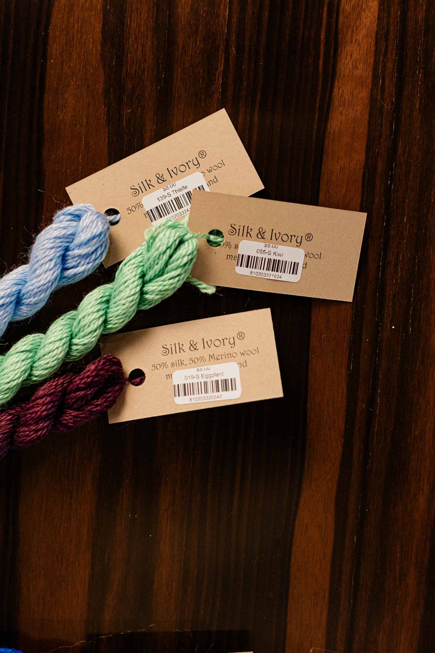 Three skeins of needlepoint yarn in blue, green, and burgundy with 'Silk & Ivory' tags on a wooden surface. | The Pepper Pot, Barrington, IL