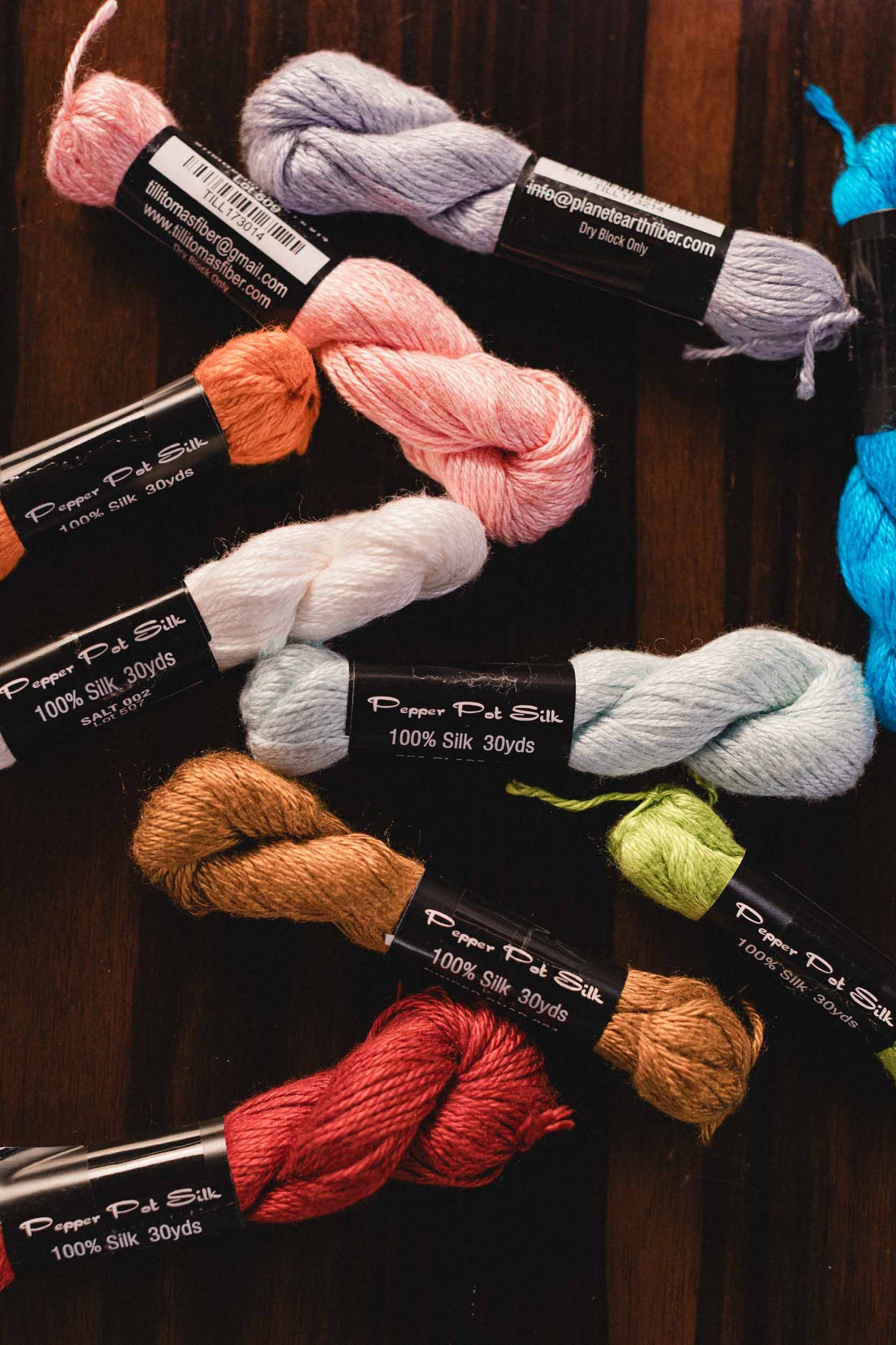 Assorted skeins of yarn by brand Pepper Pot Silk, with label that reads '100% silk 30 yards', on wooden background. | The Pepper Pot, Needlepoint and Mahjong Specialty Store | Barrington, IL