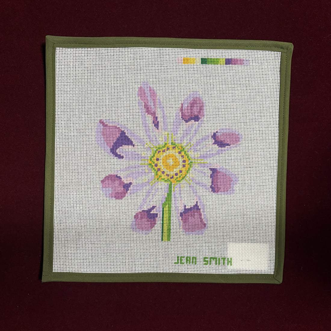 Needlepoint canvas flower design on fabric with 'Jera Smith' branding on a dark red background