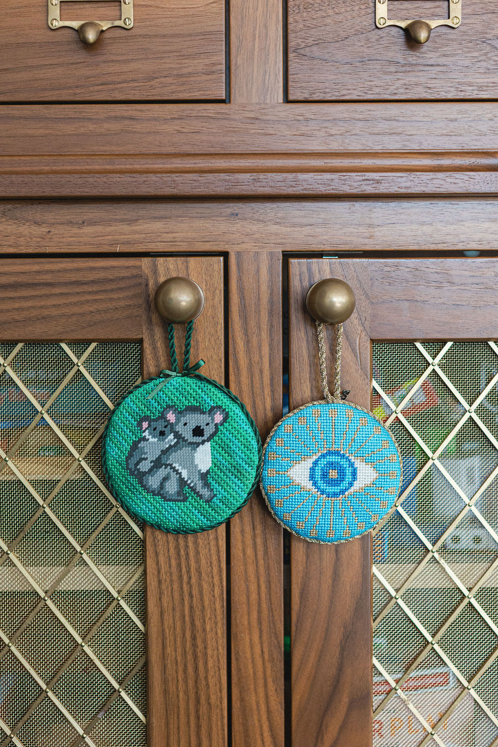 Two decorative needlepoint items, one with a koala and the other with an eye design, hanging on wooden cabinets.  | The Pepper Pot Barrington, IL 