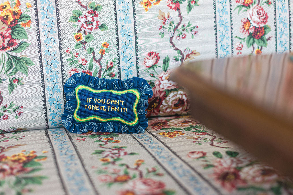 Floral-patterned needlepoint or cross-stitched fabric with a blue pillow featuring text and a brown table in the corner.  | The Pepper Pot Barrington, IL 