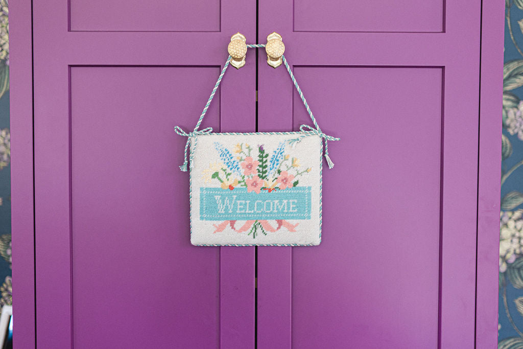 Decorative 'Welcome' needlepoint sign with floral design hanging on a purple door. | The Pepper Pot, Barrington, IL