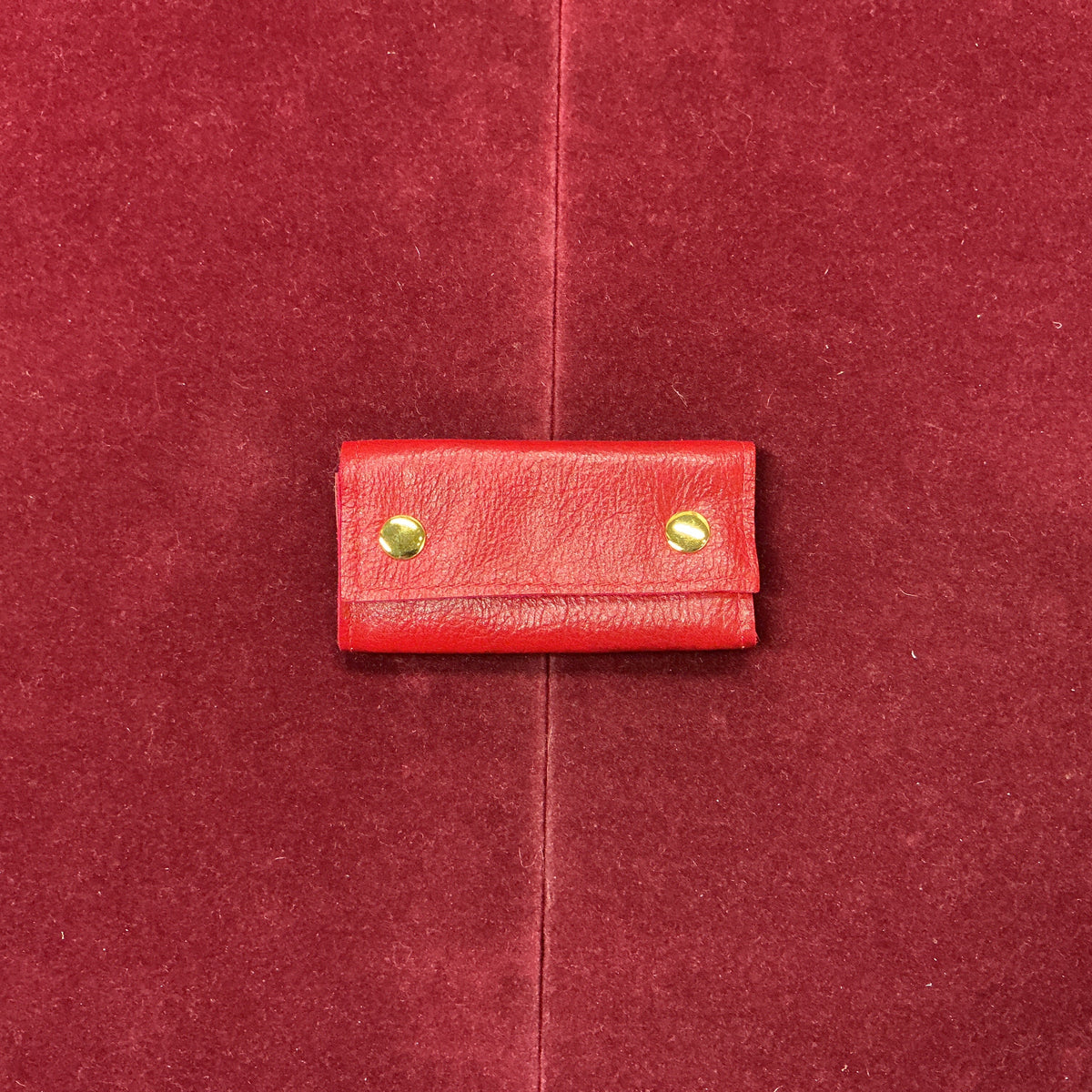 A small red leather pouch with two gold-tone metal rivets, centered on a textured red fabric background.