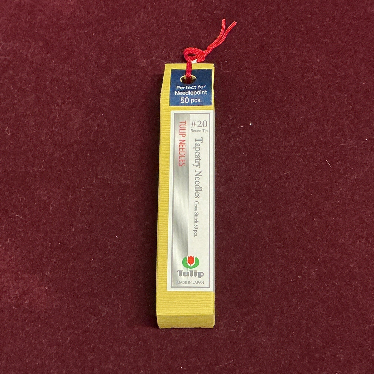A yellow rectangular box with a white label and a small red hanging string, photographed upright on a dark red textured surface.