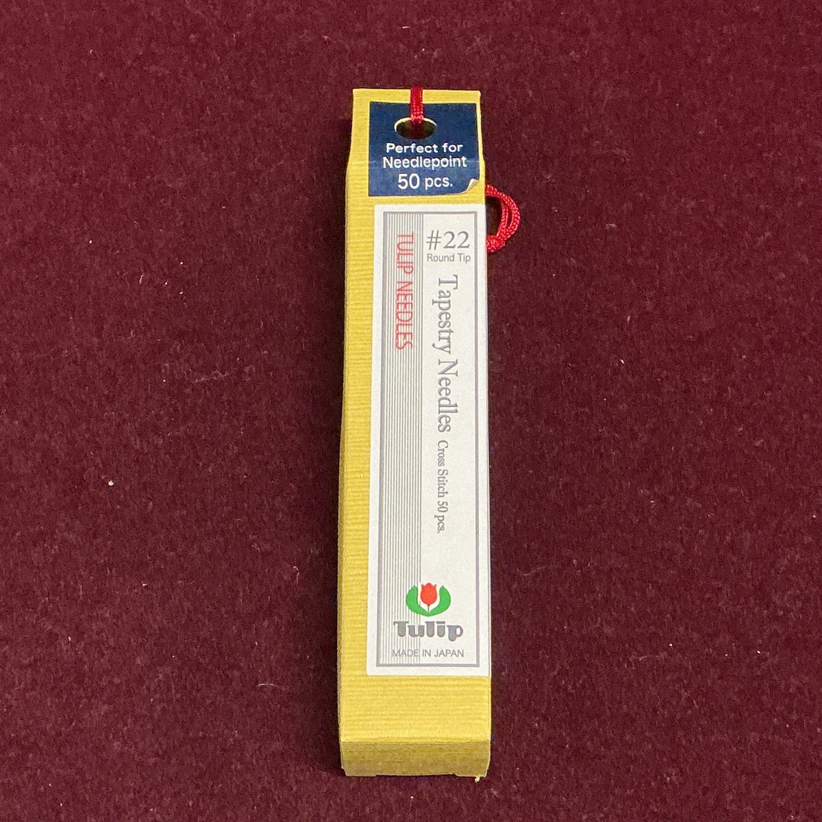 A yellow box of Tulip #22 Round Tip Tapestry Needles, 50 pieces, shown on a textured dark red background.